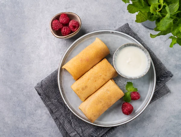 Homemade crepes with raspberry and sour cream on a plate on a light background with fresh mint close up. Healthy food concept. Top view and copy space.