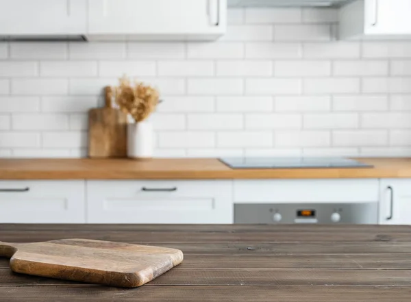 Dark wooden countertop with cutting board and free space for mounting a product or layout against the background of a blurred white kitchen. Horizontal orientation.