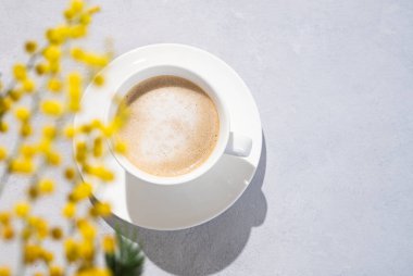 Kapuçino ya da sütlü köpüklü latte beyaz bir fonda sarı mimoza çiçekleriyle birlikte. Üst görünüm ve kopyalama alanı.