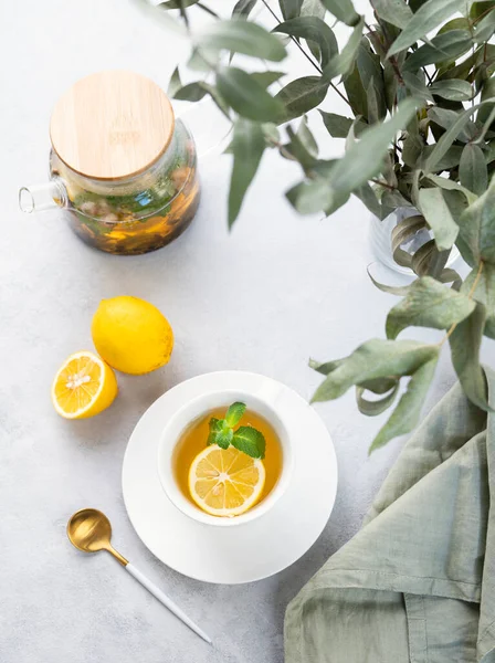 Herbal tea with lemon and mint in a white cup on a wooden plate on a light background with eucalyptus branches and a teapot. The concept of a healthy and delicious drink for   immunity. Top view.