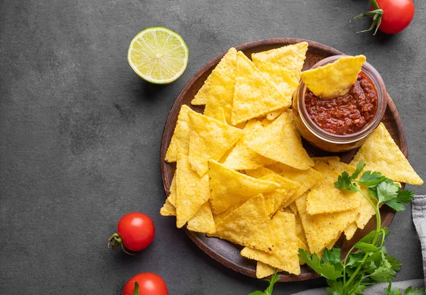 stock image Nachos or tortilla with spicy tomato sauce on a plate with fresh vegetables and herbs on a dark background. Corn chips with salsa sauce. Classic Mexican snack. Top view and copy space.