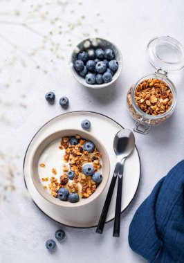 Sağlıklı bir yoğurt, tahıl gevreği ve mavi arka planda çiçekli taze yaban mersini kahvaltısı konsepti. Ev yapımı diyet ve protein yiyeceği. Üst görünüm ve kopyalama alanı.