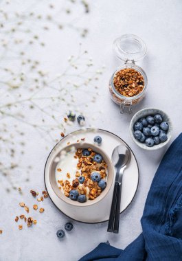 Sağlıklı bir yoğurt, tahıl gevreği ve mavi arka planda çiçekli taze yaban mersini kahvaltısı konsepti. Ev yapımı diyet ve protein yiyeceği. Üst görünüm ve kopyalama alanı.