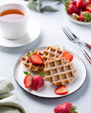 Taze çilekli ev yapımı waffle 'lar ve bir fincan çayla birlikte arka planda hafif bir tabakta pudra şekeri. Geleneksel Belçika waffle 'ları. Sağlıklı kahvaltı..