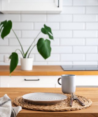 Kahvaltı için tabağı ve fincanı olan ahşap masa. Sabah ışığında İskandinav tarzı kesme tahtası ve canavar bitkisi olan bulanık beyaz mutfağa karşı.