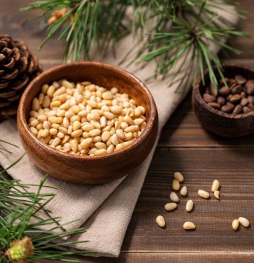 Bir kase içinde çam fıstığı (sedir) ve kahverengi ahşap bir arka planda bir avuç soyulmamış fındık ve yakın bir çam iğnesi dalında. Sağlıklı beslenme atıştırması. 