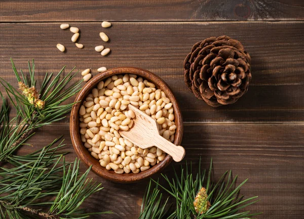 stock image Pine nuts in a bowl  on a dark wooden background with branches of pine needles and cone. The concept of a natural, organic and healthy superfood and snack. Top view.