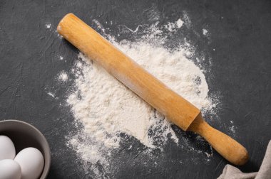 Koyu gri arka planda pişirme, saçılmış buğday unu, yumurta, merdane ve mutfak tekstili için organik malzemeler. Ev yapımı sağlıklı yiyecekler. Üst görünüm.