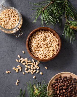 Ahşap bir kasede çam fıstığı, kavanoz ve çam iğnesi dallarıyla karanlık bir arka planda serpiştirilmiş. Doğal, organik ve sağlıklı abur cubur kavramı. Üst görünüm.