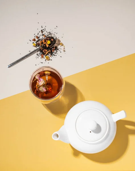 stock image Creative flat lay of a glass cup of herbal tea with rose flowers on a yellow and white background with teapot and hard shadow. Food and drink concept. Top view and copy space.