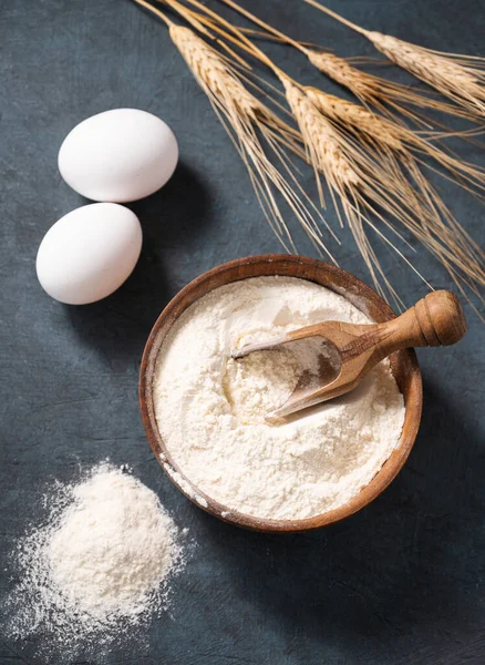 stock image Wheat flour in a wooden bowl with a scoop on a dark blue background with eggs and dry branches wheat. Organic ingredients for baking. Homemade healthy food concept.  Top view.