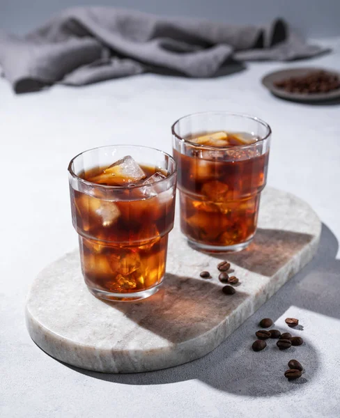 Cold brew coffee in a two glasses with ice on a marble board on a light background with coffee beans, shadows and napkin. Concept summer craft refreshing homemade drink.