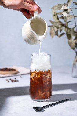 Büyük bardakta buzlu kahve, kahve çekirdekleri, dallar ve sabah gölgesi ile mavi bir fonda süt sürahisinden süt döken bir el. Yaz tatili konsepti. Ön görünüm.