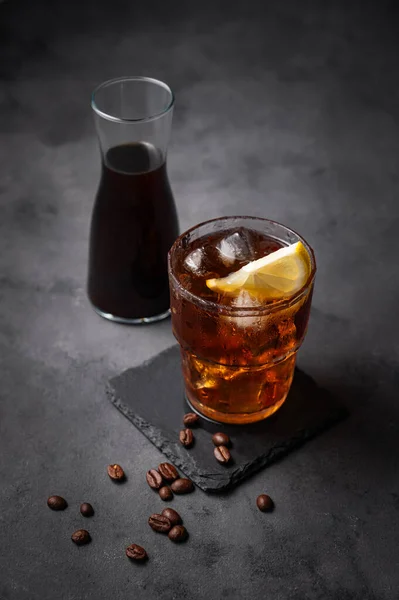 Cold brew coffee in a glass with ice and lemon on a dark concrete background with coffee beans and bottle. Concept summer craft refreshing homemade drink. Free space for text.