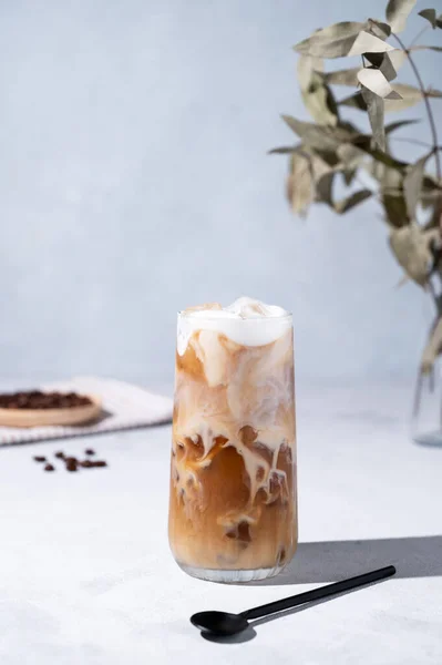 stock image Iced coffee latte in a tall glass with milk on a light background with coffee beans, spoon and morning shadows. Summer refreshment concept. Front view.