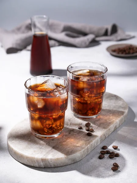 Cold brew coffee in a two glasses with ice on a marble board on a light background with coffee beans, bottle and napkin. Concept summer craft refreshing homemade drink.