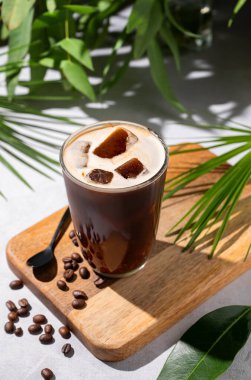 Kahve çekirdekleri ve tropikal dallarda sabah gölgeleri olan açık bir zemin üzerinde, tahta bir tahtanın üzerinde buzlu kahve bardağında demlenmiş buzlu kahve. Yaz tatili konsepti. Üst görünüm.