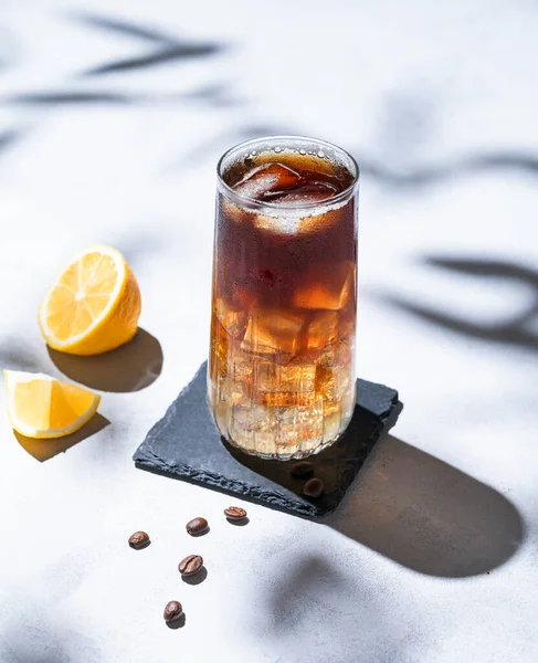 Coffee cold brew with tonic in a tall glass with ice and lemon on a light blue background with coffee beans and shadow. Trendy summer craft refreshing drink. Free space for text.