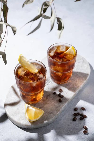 Cold brew coffee in a two glasses with lemon and ice on a marble board on a light background with coffee beans and shadows. Concept summer craft refreshing homemade drink. Top view.