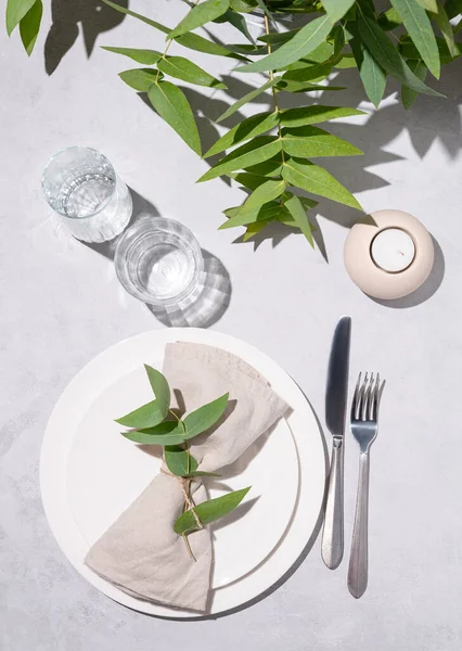 stock image Festive table setting with white plates, glasses and candle for a celebration with eucalyptus branches on a light background with shadows. Restaurant menu concept. Top view.