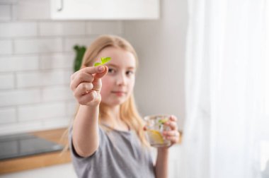 Bir kız, parlak bir mutfağın arka planında limonlu ve naneli su içer. Sağlıklı ve neşeli bir sabah konsepti.
