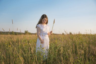 Gözlüklü genç bir kadın, tarlada hasır şapkalı beyaz bir yaz elbisesi gün batımında bir buket kır çiçeği topluyor..