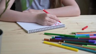 Gri tişörtlü bir kız albümde keçeli kalemle resim çiziyor, yakın planda tahta bir masada oturuyor. Yaratıcılık konsepti, hobiler, çocuk gelişimi, evde eğitim, internet dersleri.