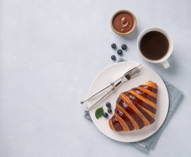 Çikolatalı ve yaban mersinli taze kruvasan beyaz bir tabakta, arka planda bir fincan sade kahveyle. Lezzetli ev yapımı kahvaltı konsepti. Üst görünüm ve kopyalama alanı. 