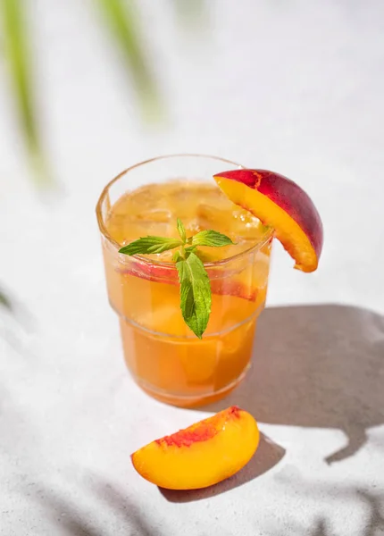 Refreshing peach tea with ice and mint. Homemade cold healthy vegetarian drink on a light background with fresh fruits, palm leaf and shadows close up.