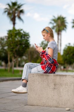 Ekose gömlekli ve kot pantolonlu bir kız güneşli bir yaz gününde elinde akıllı bir telefonla şehir parkında oturuyor. Sosyal ağlarda gençlik iletişimi.