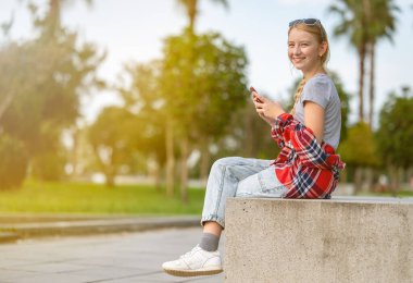 Ekose gömlekli ve kot pantolonlu bir kız güneşli bir yaz gününde elinde akıllı bir telefonla şehir parkında oturuyor. Sosyal ağlarda gençlik iletişimi.