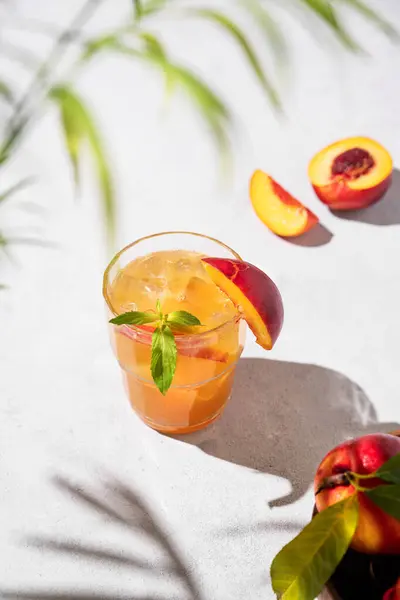 Refreshing peach tea with ice and mint. Homemade cold healthy vegetarian drink on a light background with fresh fruits, palm leaf and shadows.