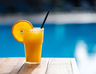 Havuzun önündeki ahşap bir masanın üstünde taze sıkılmış portakal suyu. Tatilde ferahlatıcı yaz içkisi konsepti. Metin için boş alan.