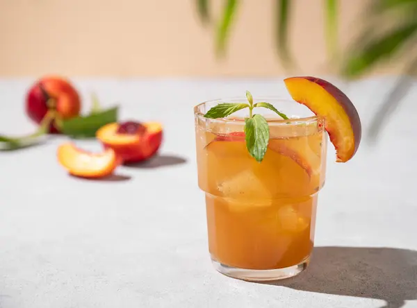 Refreshing peach tea with ice and mint. Homemade cold healthy vegetarian drink on a light background with fresh fruits, palm leaf and shadows close up. Front view and copy space.