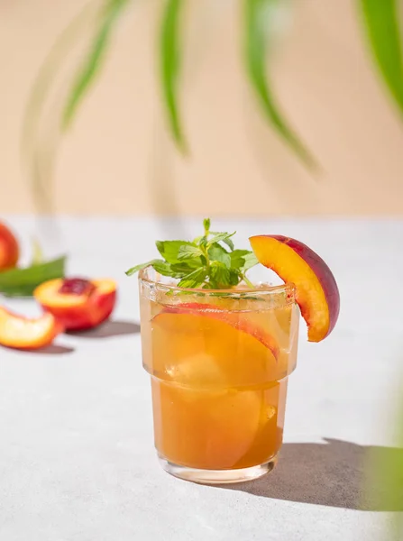 Peach tea with ice and mint. Homemade cold healthy vegetarian drink on a light background with fresh fruits, palm leaf and shadows. Front view.