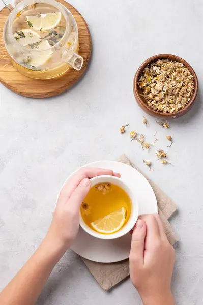 Hands holding a glass cup with chamomile herbal tea on a light background with dry flowers and teapot. The concept of a healthy drink for health and immunity. Top view.