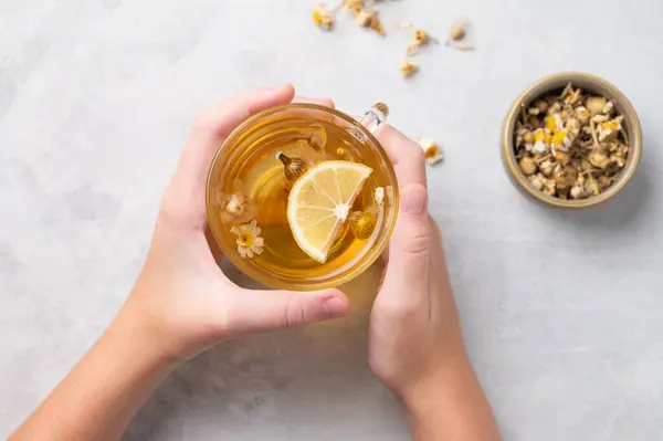 Hands holding a glass cup with chamomile herbal tea on a light background with dry flowers. The concept of a healthy drink for health and immunity. Top view.