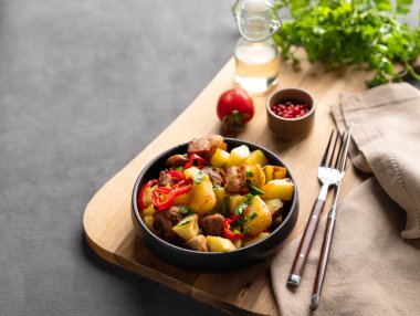 Patates, soğan ve biberli kızarmış domuz eti. Ulusal ev yapımı Gürcistan yemeği, siyah bir kâse içinde siyah bir tahta üzerinde baharatlar, taze domates ve bitkilerle birlikte arka planda. Boşluğu kopyala.