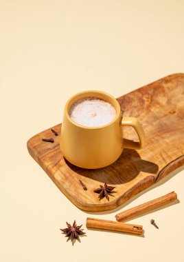 Masala latte çayı. Geleneksel Hint içeceği, baharat ve bitki karışımı. Köpüklü bir içeceği olan sarı bir kupa. Etrafı tarçın çubuklarıyla çevrili. Ve sarı arka plandaki ahşap bir tahtanın üzerinde yıldız anason.. 