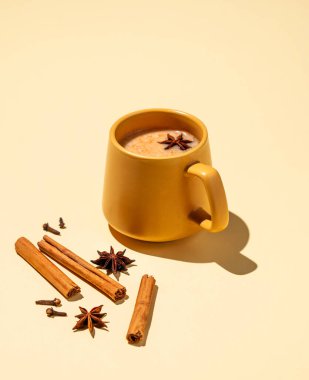Masala latte çayı. Geleneksel Hint içeceği, baharat ve bitki karışımı. Etrafı tarçın çubuklarıyla çevrili köpüklü sarı bir kupa ve sarı arka planda yıldız anason. Boşluğu kopyala.