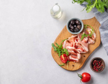 Hafif arka planda zeytin, baharat, domates ve bitkilerle birlikte domuz pastırmalı veya jambonlu düz bir tahta. Antipasto konsepti. Üst görünüm ve kopyalama alanı