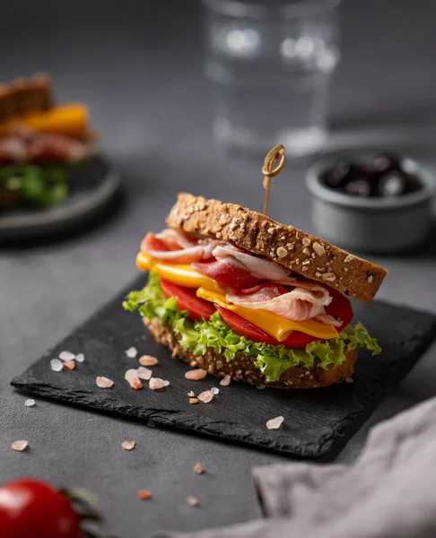 Club sandwich on a board with a  bacon, cheese, tomato and lettuce on a dark background with olives and glass of water. The concept of a healthy snack for breakfast or lunch.