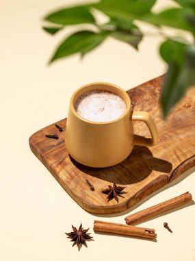 Masala çayı. Geleneksel Hint içeceği, baharat ve bitki karışımı. Köpüklü bir içeceği olan sarı bir kupa. Etrafı tarçın çubuklarıyla çevrili. Ve sarı arka plandaki ahşap bir tahtanın üzerinde yıldız anason.. 