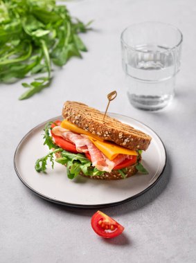 Çedar peynirli tahıllı ekmek ve domates ve roka ile doldurulmuş domuz pastırmalı tost sandviçi, bir bardak su ve bitkiyle hafif bir zemin. Sağlıklı ve beslenme kahvaltısı kavramı. 