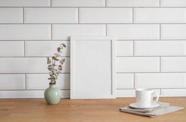 Beyaz kiremit bir duvara karşı boş bir tuval ve vazo ve fincanla ahşap bir masanın üstünde duran bir çerçeve. Fotoğraf, poster ya da resim tasarımı. Boşluğu kopyala.
