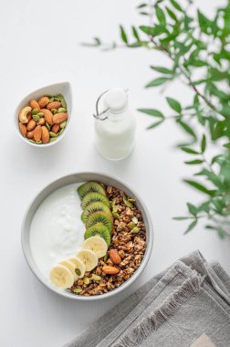 Yeşil dalı olan beyaz bir masada muzlu ve kivili granola. Düz konsept vejetaryen yoğurt, fındık, müsli ve taze meyve. Üst görünüm. 