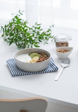 Muzlu ve kivili granola beyaz bir masada, pencerenin arka planında yeşil bir dal var. Sağlıklı kahvaltı konsepti vejetaryen yoğurt, müsli ve taze meyve..