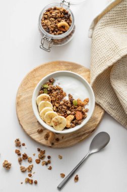 Tahtaya muz ve fındık koyarak beyaz bir masada peçete ve kaşıkla. Sağlıklı kahvaltı konsepti: Vejetaryen yoğurt, müsli ve taze meyve.. 