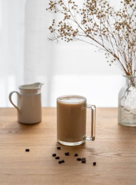 Bir bardak sütlü kahve, ahşap bir masanın üzerinde kahve çekirdekleri ve kuru buketli bir pencerenin arka planında. Sabah içeceği konsepti..