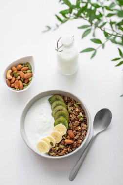 Yeşil dalı ve kaşığı olan beyaz bir masada muz ve kivi ile granola. Düz konsept vejetaryen yoğurt, fındık, müsli ve taze meyve. Üst görünüm. 